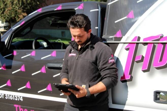 A man in a black hoodie stands next to a truck, holding a tablet and looking focused. The truck features pink graphics.